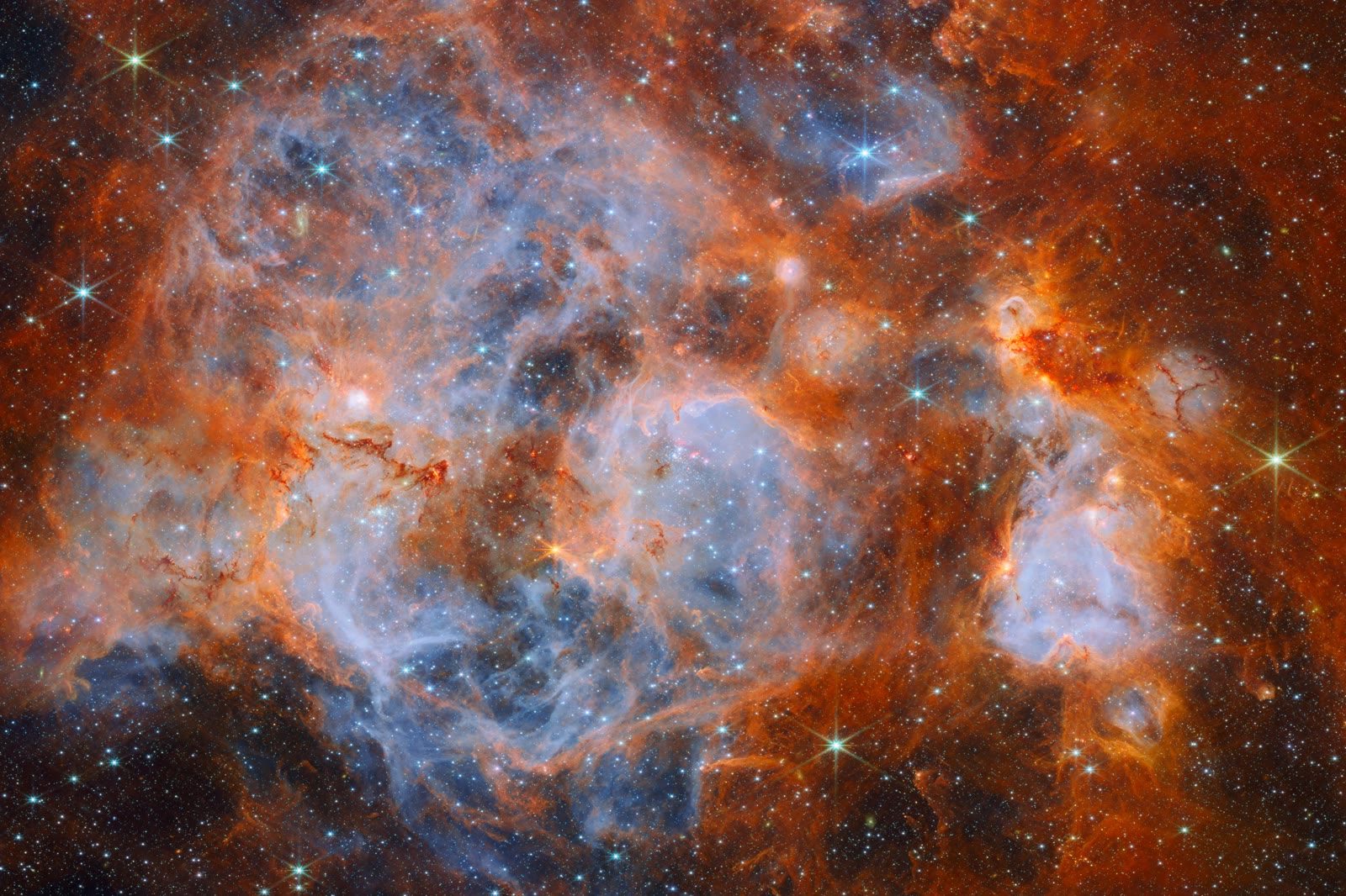 Webb near-infrared image of the nebula N159: wispy blue gas, thick orange clouds with dark ridges, thin red filaments and many stars.