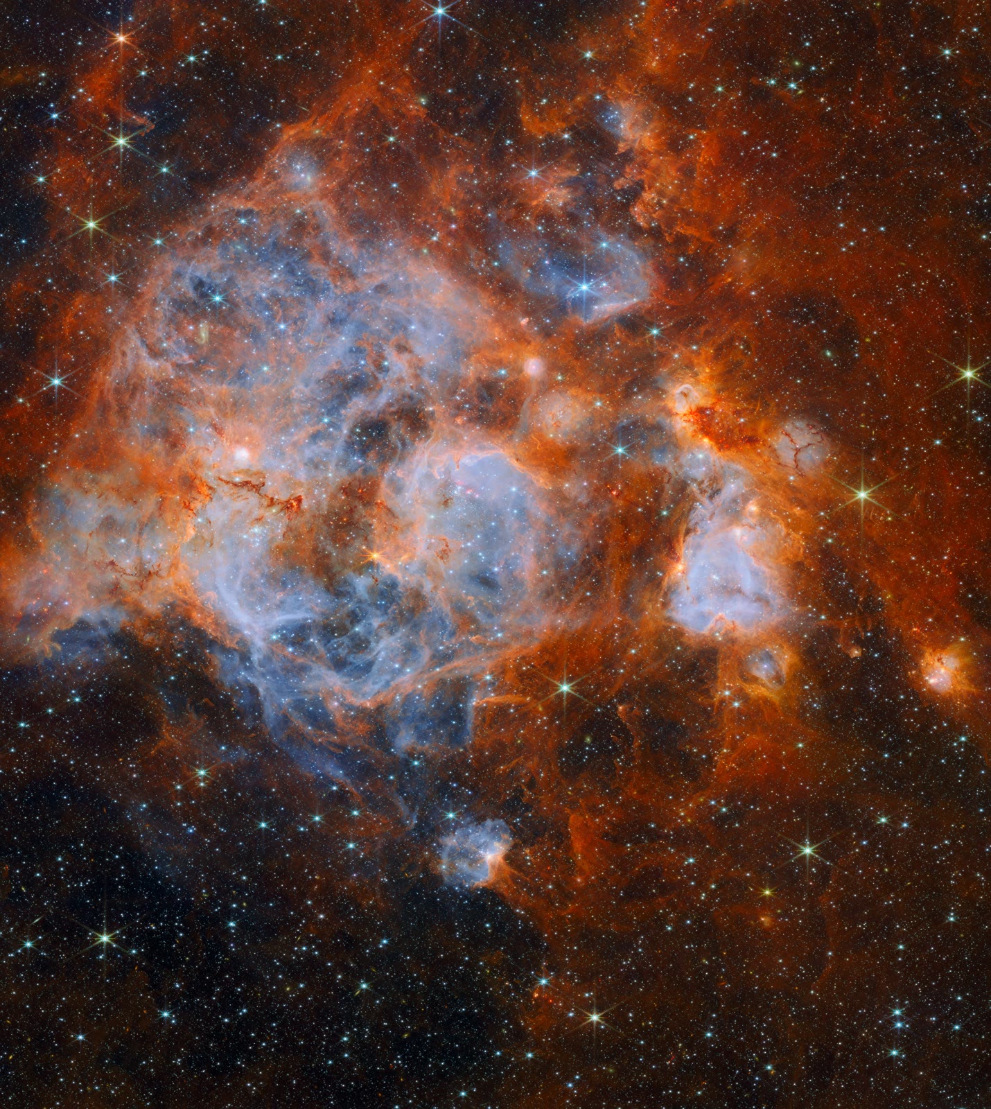 Wider Webb view of N159: the glowing nebula of blue and orange gas surrounded by a dark field of stars.